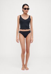 Black ribbed crop top with a scoop neckline paired with black bikini bottoms. Model wears black sunglasses, standing barefoot against a neutral background.
