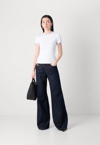 Witte t-shirt met korte mouwen en lichtblauwe bies, gecombineerd met brede, donkerblauwe denim jeans. Zwarte gestructureerde handtas in de linkerhand.