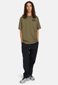 Oliven grøn bomulds t-shirt med korte ærmer, rund hals og et trykt logo på venstre bryst. Kombineret med mørkeblå bukser og sølvfarvede sneakers.