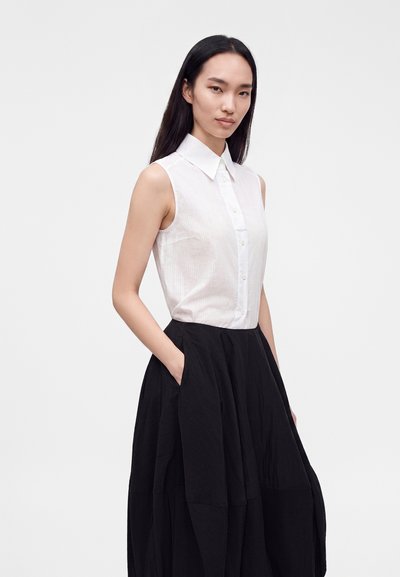 Femme aux longs cheveux foncés portant une chemise blanche sans manches à boutons et une jupe noire taille haute avec des poches, debout devant un fond uni.