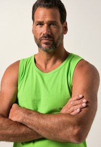 Green cotton tank top with wide shoulder straps, fitted design, and smooth texture. Features crossed arms in a neutral background.