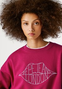 Mladá žena s objemnými kudrnatými vlasy, oblečená v magentovém svetru s bílým vyšitým logem ve tvaru rtů "Pepe Jeans London".