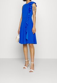 Královsky modré šaty s volánkovými detaily na rameni, těsně padnoucí korzet, rozšířená sukně a bílé sandály na vysokém podpatku. Měkká látka.