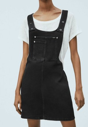 Zwarte spijkeren tuinbroekjurk met verstelbare schouderbanden, voorzak en een lichte A-lijn silhouet, gedragen over een wit T-shirt.