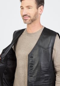 Schwarze Lederweste mit glatter Textur, ausgestattet mit einer Brusttasche und mehreren Knopfverschlüssen. Getragen über einem hellen beigefarbenen Pullover.