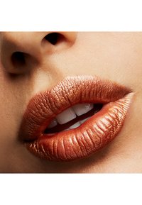 Couleur à lèvres en cuivre métallique avec une texture lisse, mettant en valeur des lèvres pleines légèrement frisées et des dents blanches en arrière-plan.