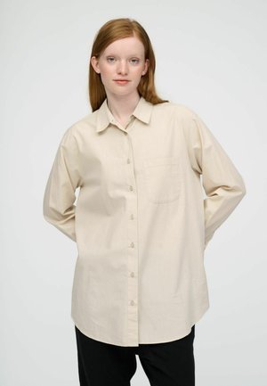 Jonge vrouw met rood haar, gekleed in een beige blouse met knopen en zwarte broek, staand met de handen achter haar rug op een witte achtergrond.