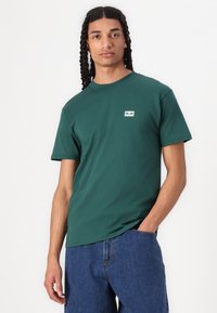 Πράσινο κοντομάνικο μπλουζάκι από βαμβάκι, με στρογγυλή λαιμόκοψη και μικρή ετικέτα λογότυπου στο στήθος. Συνδυάζεται με ανοιχτό μπλε τζιν παντελόνι.