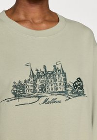 Ανοιχτό πράσινο φούτερ με λεπτομερή σχέδιο σε ναυτικό μπλε ενός αρχοντικού με δέντρα, σημαίες και το κείμενο "Mallon" κάτω από την εικόνα.