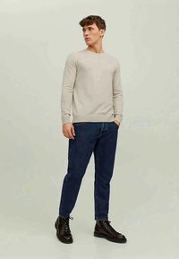 Man wearing beige long-sleeve sweater, dark blue jeans, and black lace-up boots, standing with one hand in pocket, looking to side.