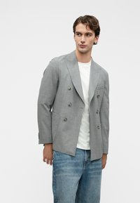 Νεαρός άνδρας φορώντας γκρι σακάκι με διπλό κουμπί πάνω από άσπρη πουκάμισο και μπλε τζιν, κοιτάζοντας δεξιά σε έναν απλό φόντο.