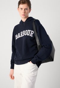 Sweatshirt azul marinho feito de mistura de algodão, apresentando "BARBOUR" em letras brancas. Combinado com calças brancas e uma bolsa tote preta.
