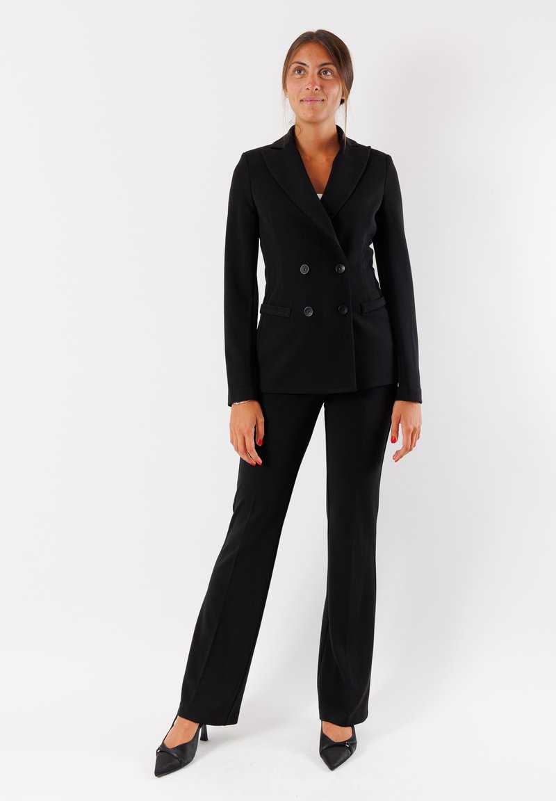 Completo elegante nero con giacca doppiopetto e rever a punte, pantaloni dritti e scarpe con tacco a punta. Tessuto dalla superficie liscia.