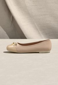 Μπεζ μπαλαρίνα με γυαλιστερό καπάκι δαχτύλου και μικρή λεπτομέρεια φιόγκου, τοποθετημένη πάνω σε ουδέτερο υφασμάτινο φόντο και ανοιχτή επιφάνεια.