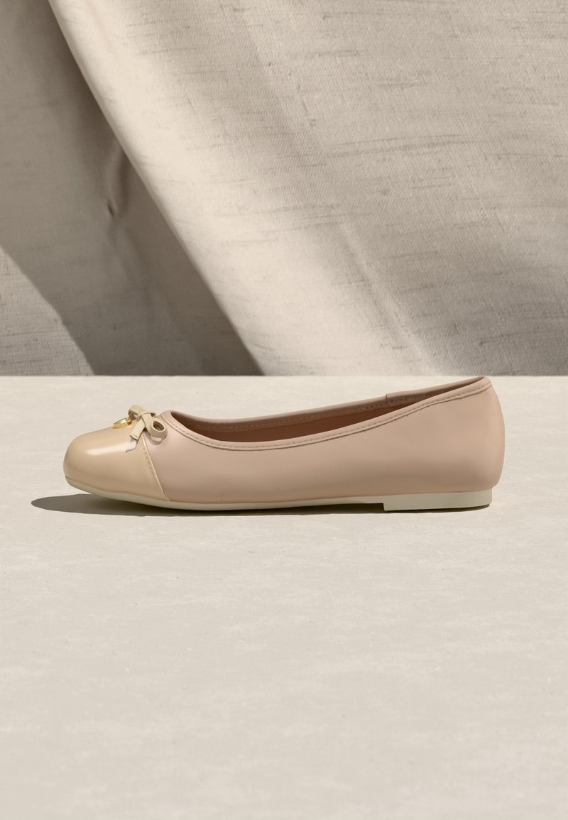 Μπεζ μπαλαρίνα με γυαλιστερό καπάκι δαχτύλου και μικρή λεπτομέρεια φιόγκου, τοποθετημένη πάνω σε ουδέτερο υφασμάτινο φόντο και ανοιχτή επιφάνεια.