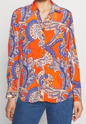 Langærmet skjorte med knapper med lyst orange, lilla og beige paisley-mønster, båret med blå jeans.