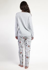Mujer de pie descalza, de espaldas, vestida con una camiseta de manga larga gris claro y pantalones de pijama con estampados de relojes, cartas y personajes de dibujos animados.