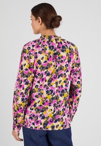 Blusa a maniche lunghe con motivo floreale in rosa, giallo e blu navy su sfondo chiaro. La parte anteriore presenta un collo alto con dettaglio arricciato.