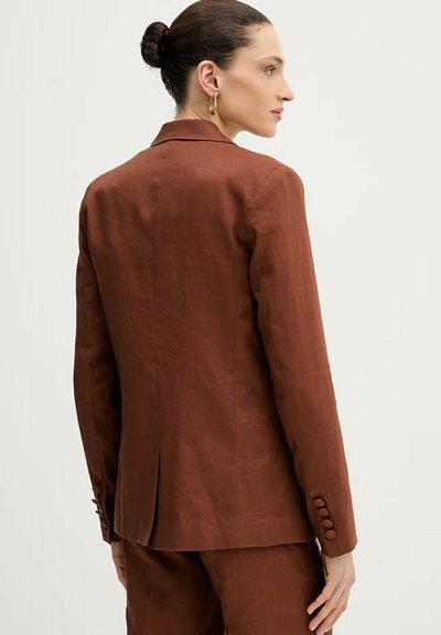 Kvinde med håret sat op i knold iført en brun skræddersyet blazer og matchende bukser, vist fra ryggen og sideprofil mod en ensfarvet baggrund.