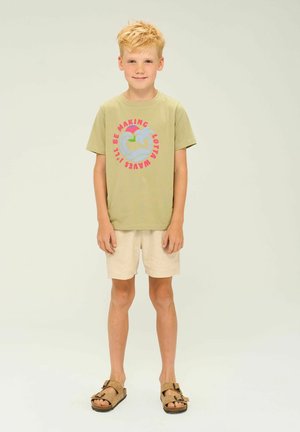 T-shirt vert clair avec un graphique de palmier coloré ; shorts en lin beige ; sandales marron à deux brides ; silhouette droite, style décontracté.