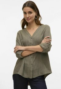 Olijfgroene button-up shirt, lichtgewicht, textuurstof, V-hals, opgerolde mouwen met knopen, gebogen zoom, gecombineerd met donkere jeans.