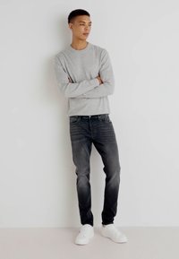 Cămașă gri cu mânecă lungă, blugi de nuanță închisă și adidași albi. Blugii au ușoare decolorări și detalii de pălmi. Un outfit simplu și casual.