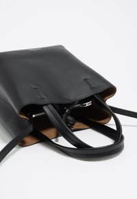 Zwarte leren tote met een gladde buitenkant, open bovenkant en korte handvatten. Het interieur heeft een ritsvak en een suede-achtige voering.