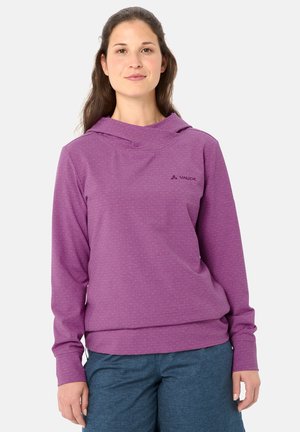 Vrouw draagt een lange mouwen paarse hoodie en blauwe broek, staat tegen een effen lichte achtergrond, kijkt naar voren met een lichte glimlach.