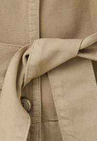 Gros plan d'une ceinture en tissu beige nouée en nœud avec des coutures visibles et un bouton métallique détaillé en dessous.