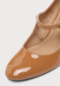 Chaussure Mary Jane en cuir verni marron clair brillant avec un bout arrondi et une fine bride avec une petite boucle dorée.