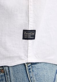 Witte linnen shirt met een zwart etiket met de tekst "Petrol Ind" genaaid aan de zoom, gecombineerd met lichtblauwe denim jeans.