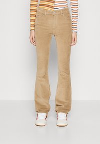 Pantalons évasés en corduroy marron avec une texture douce, poches avant et une taille avec fermeture à bouton, associés à des baskets blanches.