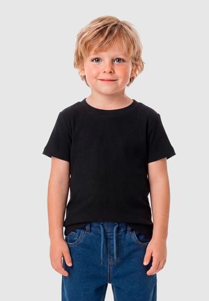 Joven rubio de ojos claros, con una camiseta negra de manga corta y jeans azules, de pie frente a un fondo claro y sencillo.