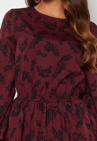 Burgundy mekko pitkillä hihoilla, rypytetty vyötärö ja musta kukkakuosi; valmistettu pehmeästä kankaasta, jolla on sileä pinta.