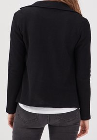Persona che indossa una giacca nera a maniche lunghe sopra una camicia bianca, abbinata a jeans grigio scuro, vista da dietro.