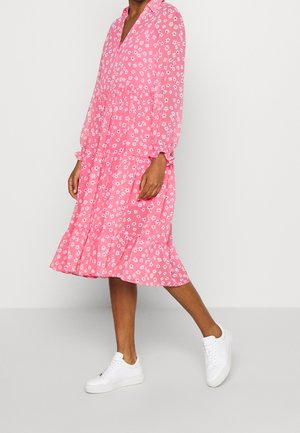 Person trägt ein rosa Midikleid mit Blumenmuster, langen Ärmeln und weißen Sneakers, steht vor einem schlichten hellen Hintergrund.