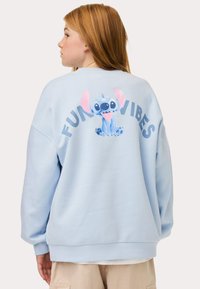 Felpa azzurra chiara con un personaggio dei cartoni animati, Stitch, e la scritta "FUN VIBES" in lettere nere in grassetto sul retro. Tessuto morbido e casual.