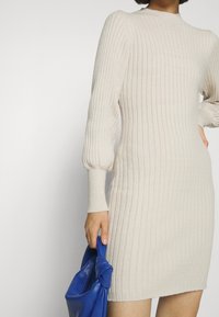 Beżowa dzianinowa sukienka z prążkowanym wzorem, z długimi bufiastymi rękawami i wysokim kołnierzem. Ma dopasowany fason i trzyma niebieską skórzaną torebkę.
