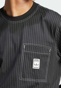 Svart randig skjorta med vita smala ränder, rund halsringning, en bröstficka och en Adidas logotyp på etiketten på fickan. Slät textur.