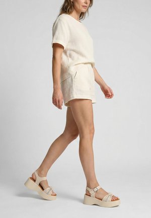 Kvinde, der går, iført cremefarvet kortærmet tekstureret top, matchende shorts og beige plateau-sandaler med ankelremme.