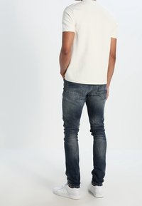 Vaalean beige lyhythihainen t-paita, slim fit, yhdistettynä tummansinisiin farkkuihin ja valkoisiin lenkkikenkiin, nähtynä takaa.