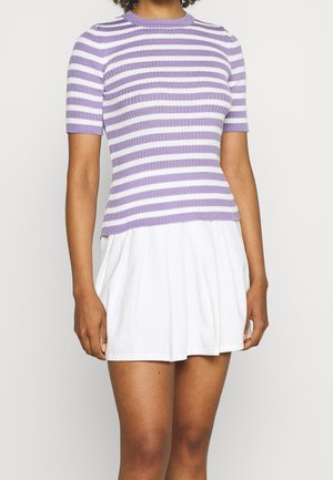 Frau trägt ein lila-weiß horizontal gestreiftes Kurzarmshirt kombiniert mit einem weißen plissierten Rock vor einfarbigem Hintergrund.