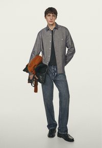 Grijze button-up shirt, zwarte t-shirt, blauwe jeans, zwarte schoenen, een oranje trui en een zwarte tas vasthoudend. Eenvoudige, casual outfit.
