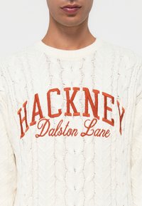 Strukturirana krema pletena jakna z izrazitim oranžnim grafikonom "HACKNEY" in "Dalston Lane" pod njim. Okrogla izrezana ovratnica s kabelnim pletenjem.