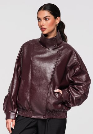 Frau mit dunkel zurückgebundenem Haar trägt eine dunkelviolette, übergroße Lederjacke mit hohem Kragen und elastischer Taille und schaut nach rechts.