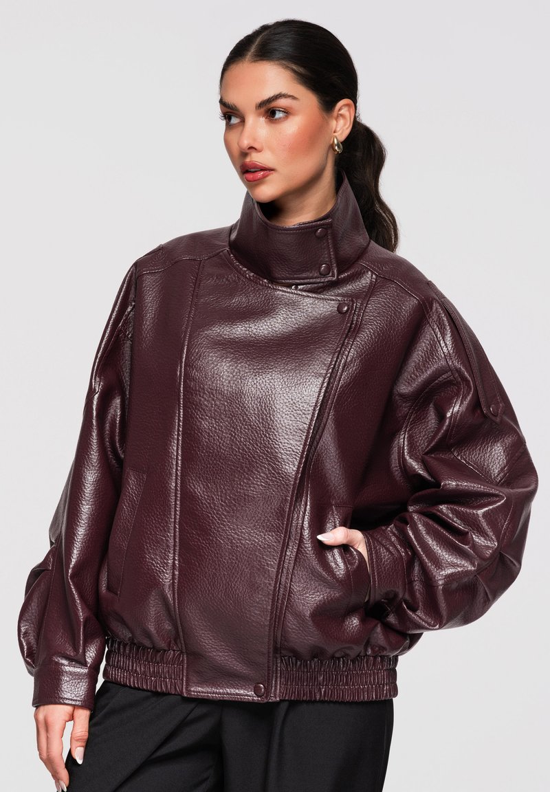 Mujer con el pelo oscuro recogido hacia atrás, lleva una chaqueta de cuero oversize de color púrpura oscuro con cuello alto y cintura elástica, mirando hacia la derecha.