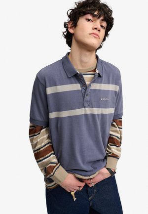 Junge Person mit lockigem Haar trägt ein blau gestreiftes Polohemd über einem langärmligen gestreiften Shirt, hält Schlüssel und trägt Ringe und eine Uhr.