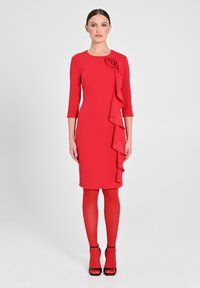 Abito rosso con scollo rotondo, maniche a tre quarti, dettaglio a volant su un lato e una rosa come decorazione; abbinato a calzamaglie rosse e tacchi neri.