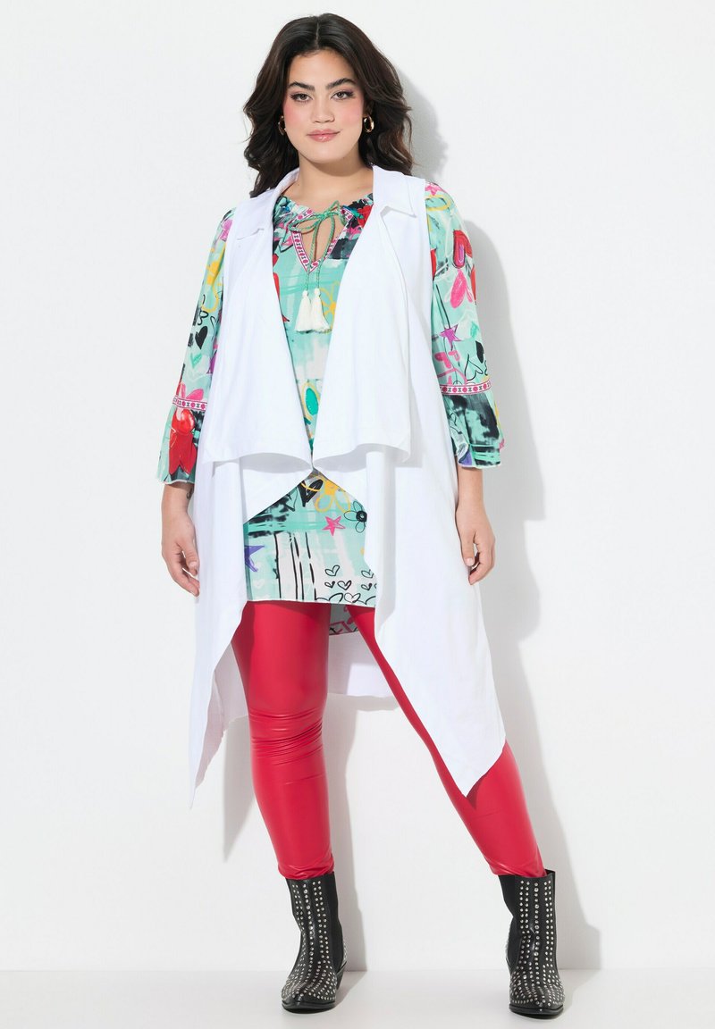 Gilet bianco senza maniche sovrapposto a una maglietta colorata con motivo e colletto a fiocco, abbinato a legging rossi e stivaletti neri con borchie.