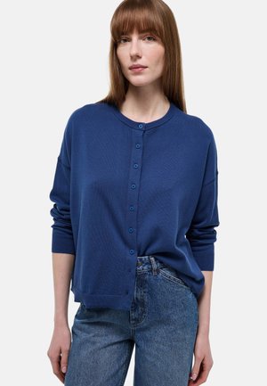 Femme aux cheveux bruns raides portant un cardigan bleu boutonné et un jean bleu, debout devant un fond clair uni.